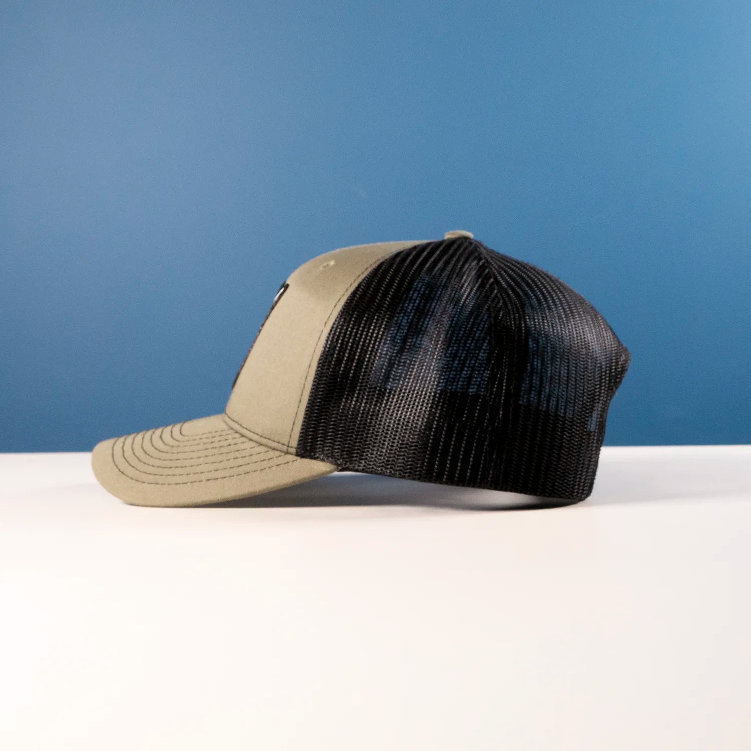 OD Green and black trucker cap on a white surface with a blue background